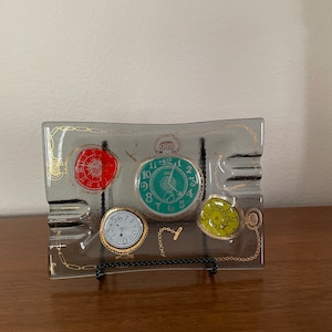 May include: A glass ashtray with a decorative design of four clocks with gold chains. The clocks are in different colors, including red, green, and yellow.
