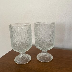 May include: Two clear, textured glass goblets with a stem and a wide, cylindrical body. The glass has a rough, bark-like texture. The goblets are displayed on a wooden surface. Ideal for serving beverages.