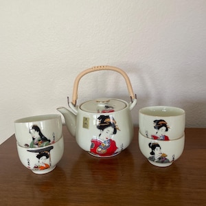 May include: A white ceramic tea set featuring a teapot with a woven handle and lid, and four matching teacups. Each piece is decorated with a geisha design in red, black, and gold. The set is displayed on a wooden surface.