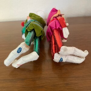 May include: Two vintage felt Christmas elf ornaments with metallic green and red legs and white boots. The ornaments have a blue tag that reads "All New Materials Made in Japan".