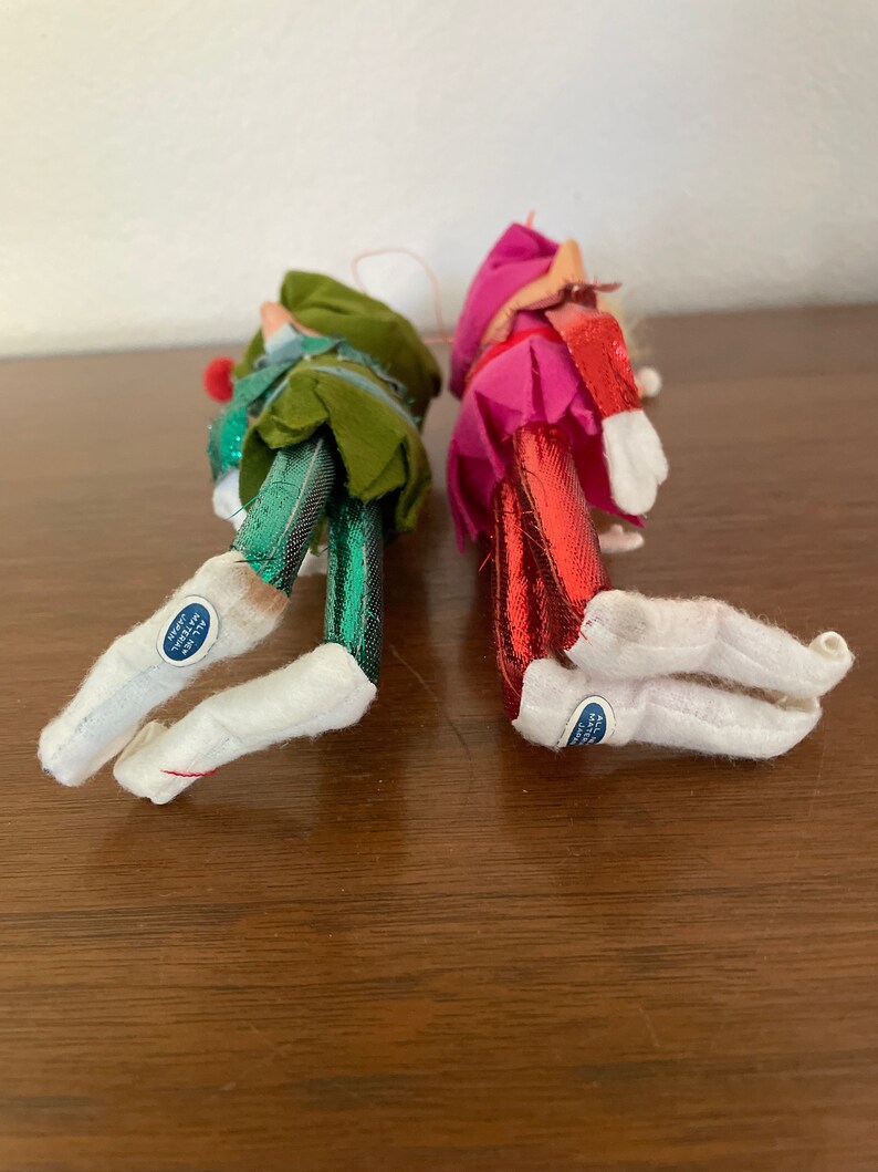 May include: Two vintage felt Christmas elf ornaments with metallic green and red legs and white boots. The ornaments have a blue tag that reads "All New Materials Made in Japan".