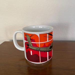 May include: White ceramic coffee mug with a wraparound design featuring a stylized airplane against a red and orange sunset background. The mug has a curved handle and a glossy finish. The design includes a dark brown airplane.