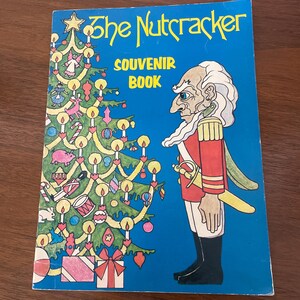 May include: A vintage souvenir book titled "The Nutcracker" with a blue cover. The cover features a colorful illustration of a decorated Christmas tree and a soldier character. The text "SOUVENIR BOOK" is also displayed.