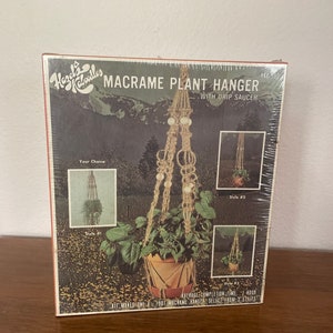May include: A box for a macrame plant hanger kit. The box shows a brown macrame plant hanger with white beads. The text on the box reads "Macrame Plant Hanger with Drip Saucer".