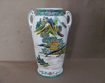May include: A white ceramic vase with two handles, featuring a hand-painted design of a Japanese landscape with mountains, trees, and a pagoda. The vase has a green and white floral pattern around the base.