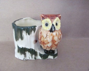 May include: A ceramic owl planter with a brown, yellow, and black color scheme. The owl is perched on a tree stump planter with green and white paint.