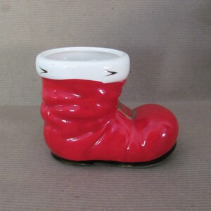 May include: A ceramic Santa boot-shaped planter. The boot is a glossy red with a white cuff and black accents. The interior is white, and the base is black. A festive holiday decoration.