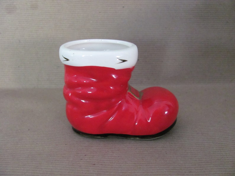 May include: A ceramic Santa boot-shaped planter. The boot is a glossy red with a white cuff and black accents. The interior is white, and the base is black. A festive holiday decoration.