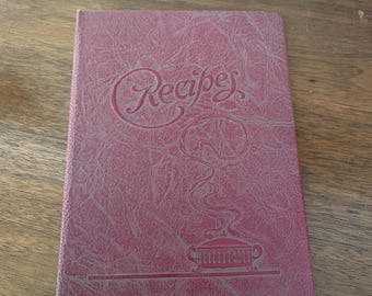 May include: A burgundy-colored recipe book with the word "Recipes" embossed on the cover. The cover also features an embossed illustration of a pot with steam rising from it. The book is rectangular and appears to be closed.