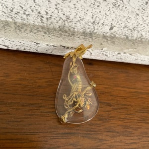 May include: A gold pendant with a clear glass teardrop shape. Inside the glass is a gold figure of a person with a red gem on their head.
