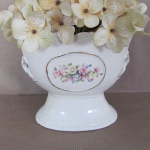 Puede incluir: Jarr&oacute;n de cer&aacute;mica blanca con un dise&ntilde;o floral y un borde festoneado. El jarr&oacute;n est&aacute; decorado con un borde dorado y presenta un dise&ntilde;o floral en relieve. El jarr&oacute;n est&aacute; lleno de flores blancas.