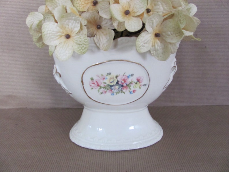 Puede incluir: Jarr&oacute;n de cer&aacute;mica blanca con un dise&ntilde;o floral y un borde festoneado. El jarr&oacute;n est&aacute; decorado con un borde dorado y presenta un dise&ntilde;o floral en relieve. El jarr&oacute;n est&aacute; lleno de flores blancas.