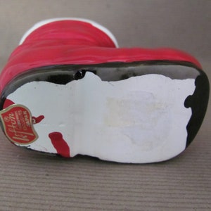 May include: A ceramic Santa boot figurine, primarily red with a white cuff and black base. The boot has a glossy finish and a small, red and gold sticker that reads "Lefton Trademark Exclusives Japan."