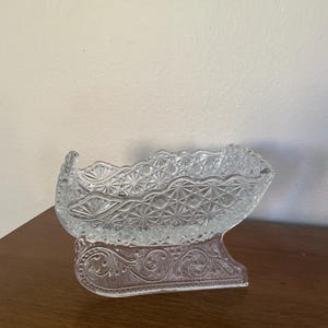 May include: Clear glass sleigh-shaped dish with a decorative pattern. The dish has a detailed, textured design and sits on a base with an ornate, etched pattern. The sleigh is made of clear glass.