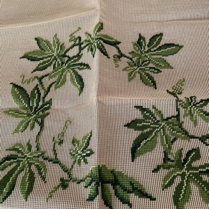 May include: A beige needlepoint canvas featuring a partially completed cross-stitch pattern of green vine leaves arranged in a circular wreath shape. The design uses various shades of green thread to create depth and detail on the grid fabric.