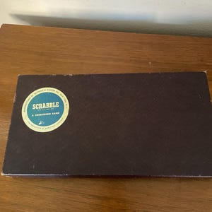 May include: A rectangular, dark brown Scrabble game box with a circular label. The label reads "SCRABBLE A CROSSWORD GAME" and includes the manufacturer's name. The box is on a wooden surface.