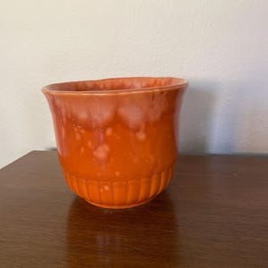 May include: A small, orange ceramic planter with a white glaze. The planter has a rounded bottom and a flared top.