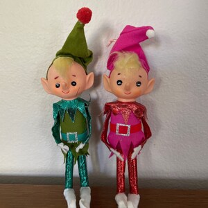 May include: Two vintage Christmas elves with felt bodies and shiny metallic legs. One elf is wearing a green hat and a green and teal outfit. The other elf is wearing a pink hat and a pink and red outfit.