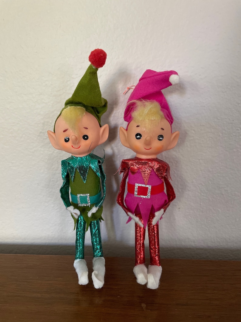 May include: Two vintage Christmas elves with felt bodies and shiny metallic legs. One elf is wearing a green hat and a green and teal outfit. The other elf is wearing a pink hat and a pink and red outfit.