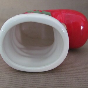 May include: A ceramic tomato-shaped container with a white oval opening. The container is primarily red with green and red accents at the top. The interior of the container is white.