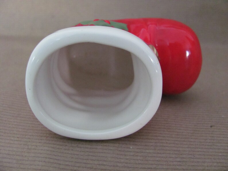 May include: A ceramic tomato-shaped container with a white oval opening. The container is primarily red with green and red accents at the top. The interior of the container is white.