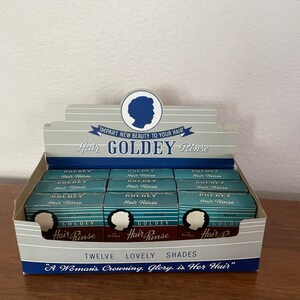 May include: A vintage display box of Goldey Hair Rinse in twelve shades. The box is white with blue and red accents. The text on the box reads "IMPART NEW BEAUTY TO YOUR HAIR" and "Hair GOLDEY Rinse". The box contains twelve small boxes of hair rinse, each with a different shade. The shades include Ash Blonde, Platinum, and Black.