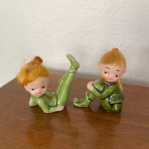 May include: Two ceramic figurines of elves. One is lying down with a leg raised, and the other is sitting with its knees bent. Both elves wear green outfits and have orange hair. The figurines are on a wooden surface.