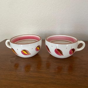 May include: Two white ceramic soup cups with handles, each adorned with a repeating pattern of stylized red and yellow leaves. The rims and interiors of the cups are a deep pink, creating a contrasting color scheme. The cups are set on a dark wooden surface.