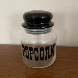 May include: A clear glass popcorn jar with a black lid. The word "POPCORN" is printed in bold, black letters on the front of the jar. The jar is sitting on a wooden surface.