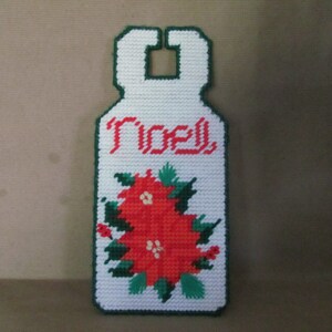 May include: White plastic door hanger with a green border. It features a red poinsettia with green leaves and the word "Noel" in red.