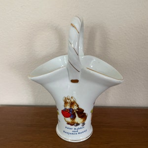 May include: White porcelain basket with a gold handle and a depiction of Peter Rabbit and Benjamin Bunny. The basket has a scalloped rim and a gold trim.