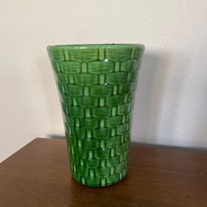 May include: A green ceramic vase with a woven basket design. The vase is cylindrical, with a slightly flared top. The glossy finish reflects light, highlighting the texture of the basket weave pattern. The vase is approximately 7 inches tall.