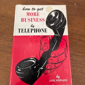 May include: A hardcover book titled How to Get More Business by Telephone by Jack Schwartz. The cover features a graphic illustration of a hand holding a black telephone receiver against a red and white background with bold black and red text.