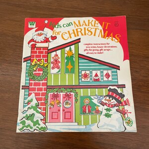 Peut inclure: Un livre vintage intitul&eacute; "Kids can MAKE IT for CHRISTMAS" avec des illustrations d'un bonhomme de neige, d'un sapin de No&euml;l et d'une maison avec une chemin&eacute;e. Le livre contient des instructions pour la fabrication de d&eacute;corations de No&euml;l, de cadeaux et de papier d'emballage.