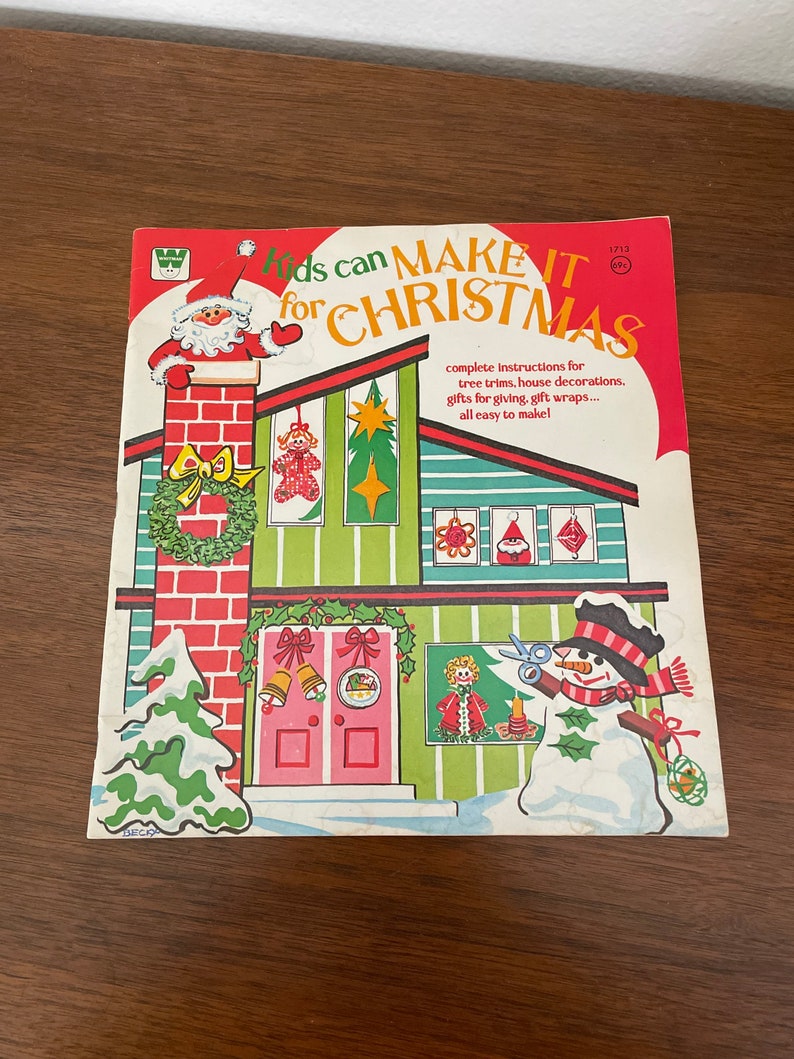 Peut inclure: Un livre vintage intitul&eacute; "Kids can MAKE IT for CHRISTMAS" avec des illustrations d'un bonhomme de neige, d'un sapin de No&euml;l et d'une maison avec une chemin&eacute;e. Le livre contient des instructions pour la fabrication de d&eacute;corations de No&euml;l, de cadeaux et de papier d'emballage.