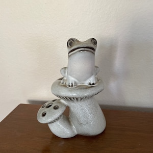 May include: A ceramic frog figurine sitting on top of a mushroom-shaped vase with a smaller mushroom-shaped spout. The frog and vase are both white with brown accents.