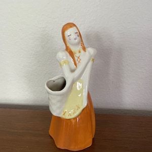 May include: A ceramic figurine of a woman with orange hair and a yellow and orange dress. She is holding a white bag in her left hand and has a closed-eye smile. The figurine is a vintage planter.