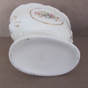 Puede incluir: Jarr&oacute;n de cer&aacute;mica blanca con un dise&ntilde;o floral y detalles dorados. El jarr&oacute;n tiene una base redonda y una abertura ancha.