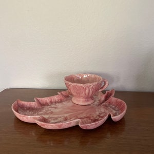 Pu&ograve; includere: Un vassoio da portata in ceramica a forma di foglia rosa con una tazza rosa abbinata. Il vassoio ha un design marmorizzato e la tazza ha un bordo smerlato.