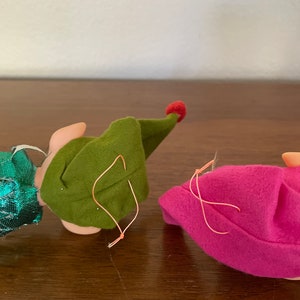 May include: Two felt elf ornaments, one green and one pink, with red felt hats. Both have a loop for hanging.