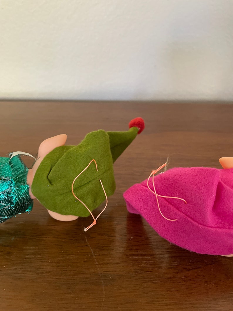 May include: Two felt elf ornaments, one green and one pink, with red felt hats. Both have a loop for hanging.