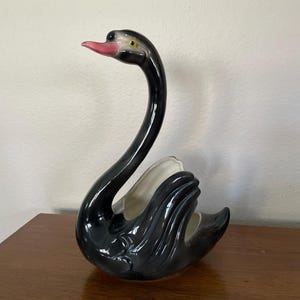 May include: A black ceramic swan planter with a pink beak and yellow eye. The swan's neck is curved and the body is hollowed out to hold a plant.