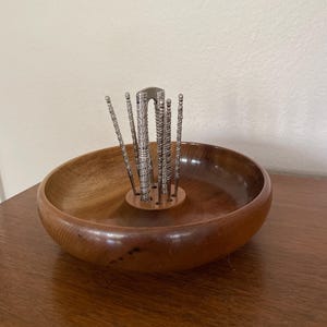 May include: A round, polished wooden bowl featuring a central circular stand with multiple holes. Inserted into these holes are several silver-toned metal nut picks and a nutcracker, all displaying intricate, textured patterns along their handles.