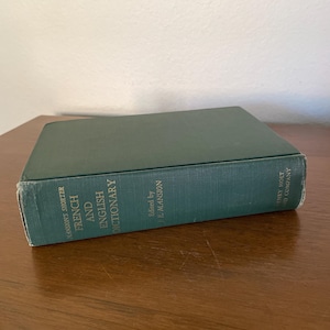 May include: A hardcover book titled "Mansion's Shorter French and English Dictionary" in a teal cover. The book is edited by J.E. Mansion and published by Henry Holt and Company. The book is resting on a wooden surface.