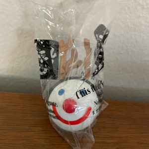 May include: A white plastic toy with a smiling face, blue eyes, a red nose, and a red mouth. The toy is packaged in a clear plastic bag with the text "This item is not a toy. Adult supervision required." printed on it.