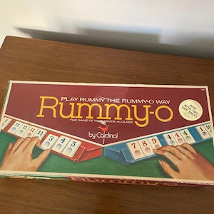 May include: A vintage Rummy-O board game box. The box is primarily burgundy with the game's title in large yellow letters. The game includes two-tier racks and is by Cardinal. The box shows hands holding game pieces.