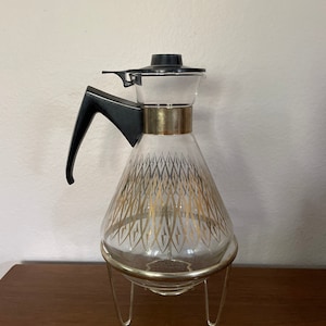 May include: A clear glass coffee pot with a gold geometric pattern and a black lid. The pot is on a gold metal stand with three legs.