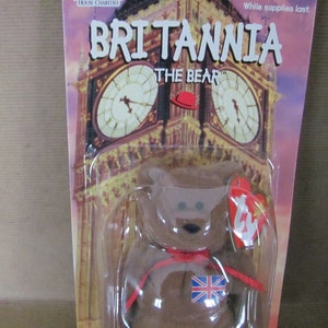 May include: A brown Beanie Baby bear named Britannia The Bear, wearing a red ribbon and a Union Jack patch. The bear is packaged in a plastic bag with a red, white, and blue background featuring a picture of Big Ben.