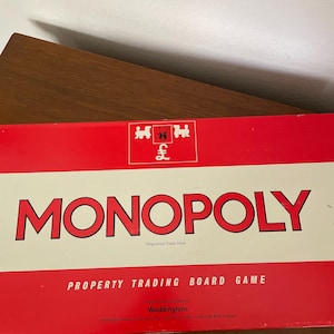 May include: A rectangular Monopoly property trading board game box with a red and white design. The box features the Monopoly logo in large red letters, a pound sterling symbol, and the text Manufactured in England by Waddingtons.