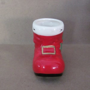 May include: A ceramic Santa boot-shaped container. The boot is primarily red with a white cuff and a gold-colored buckle. Green accents are visible on the side. The container has a black base and is designed for decorative or functional use.